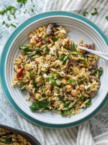 A turquoise plate with a white bowl on it filled with orzo salad sitting on a striped towel