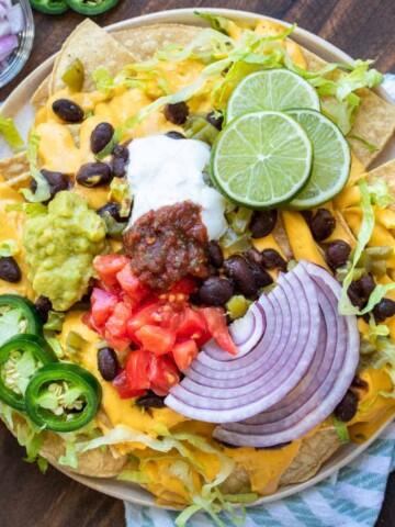 A plate of nachos with all the fixings on a striped towel with bowls of ingredients around it.