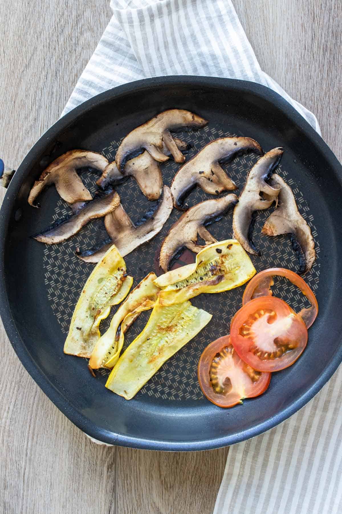 A pan with sliced mushrooms, squash and tomatoes being cooked inside