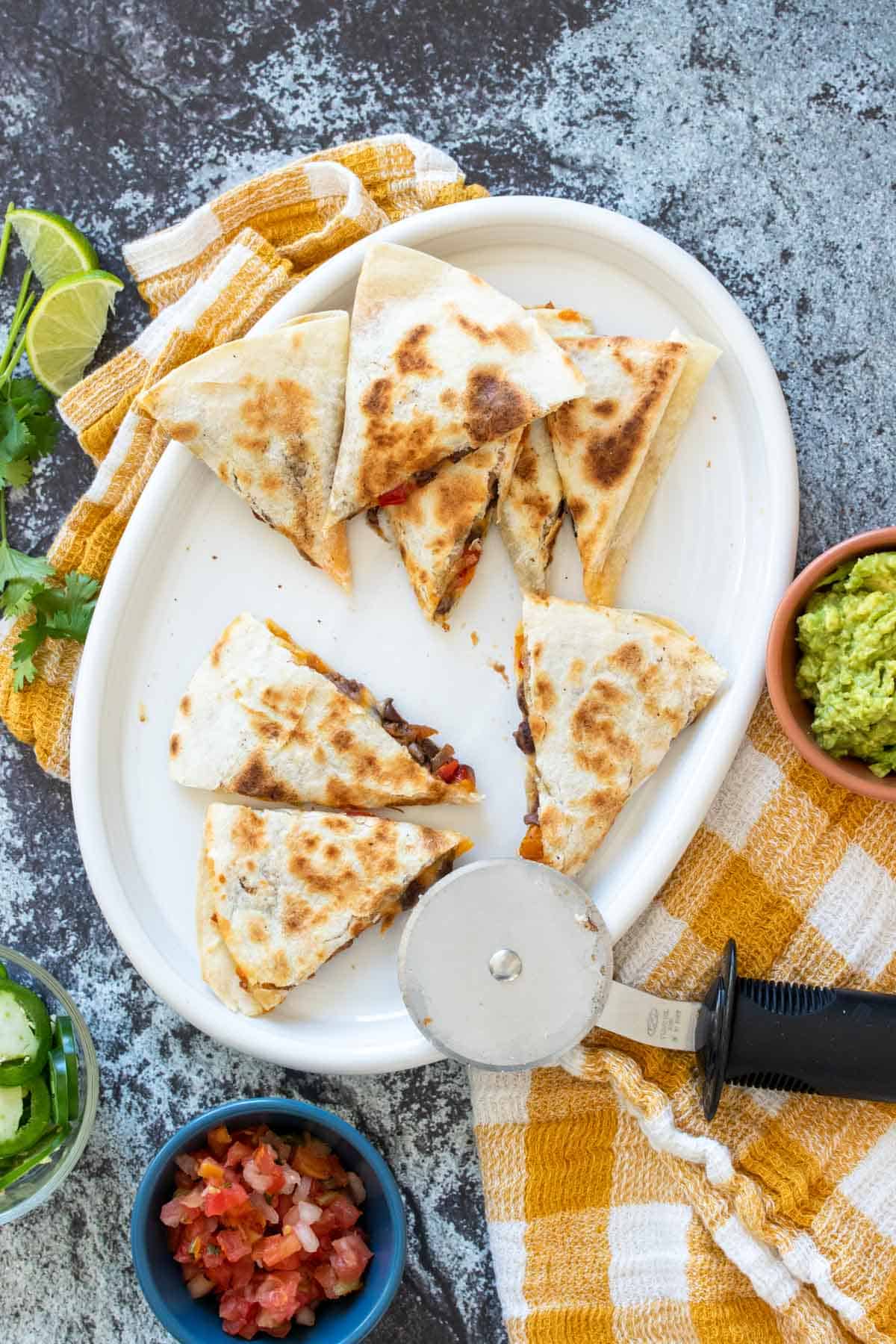 A pizza cutter next to triangles of cut quesadillas on a white plate sitting on a yellow checkered towel