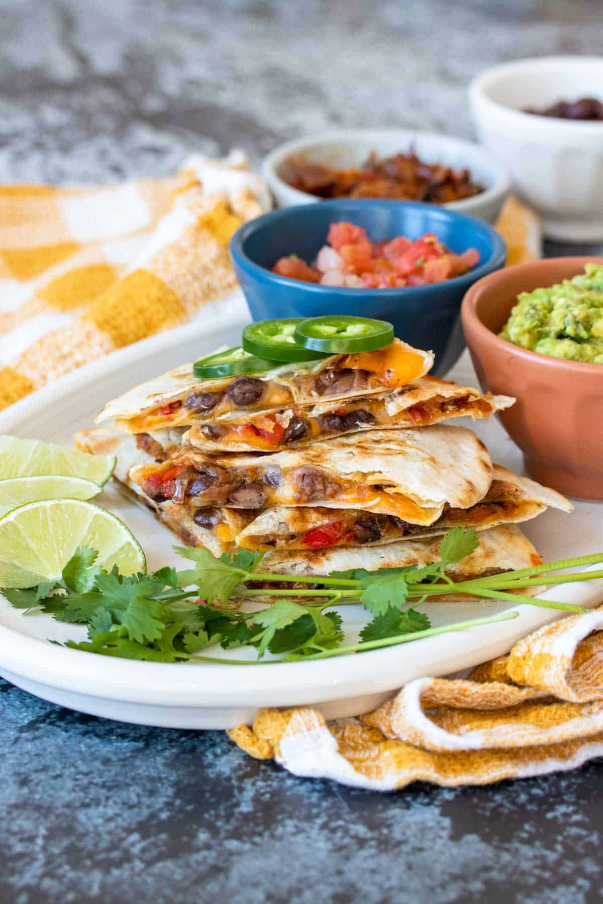 A white platter with colorful bowls of toppings and a stack of quesadillas with black beans and veggies inside