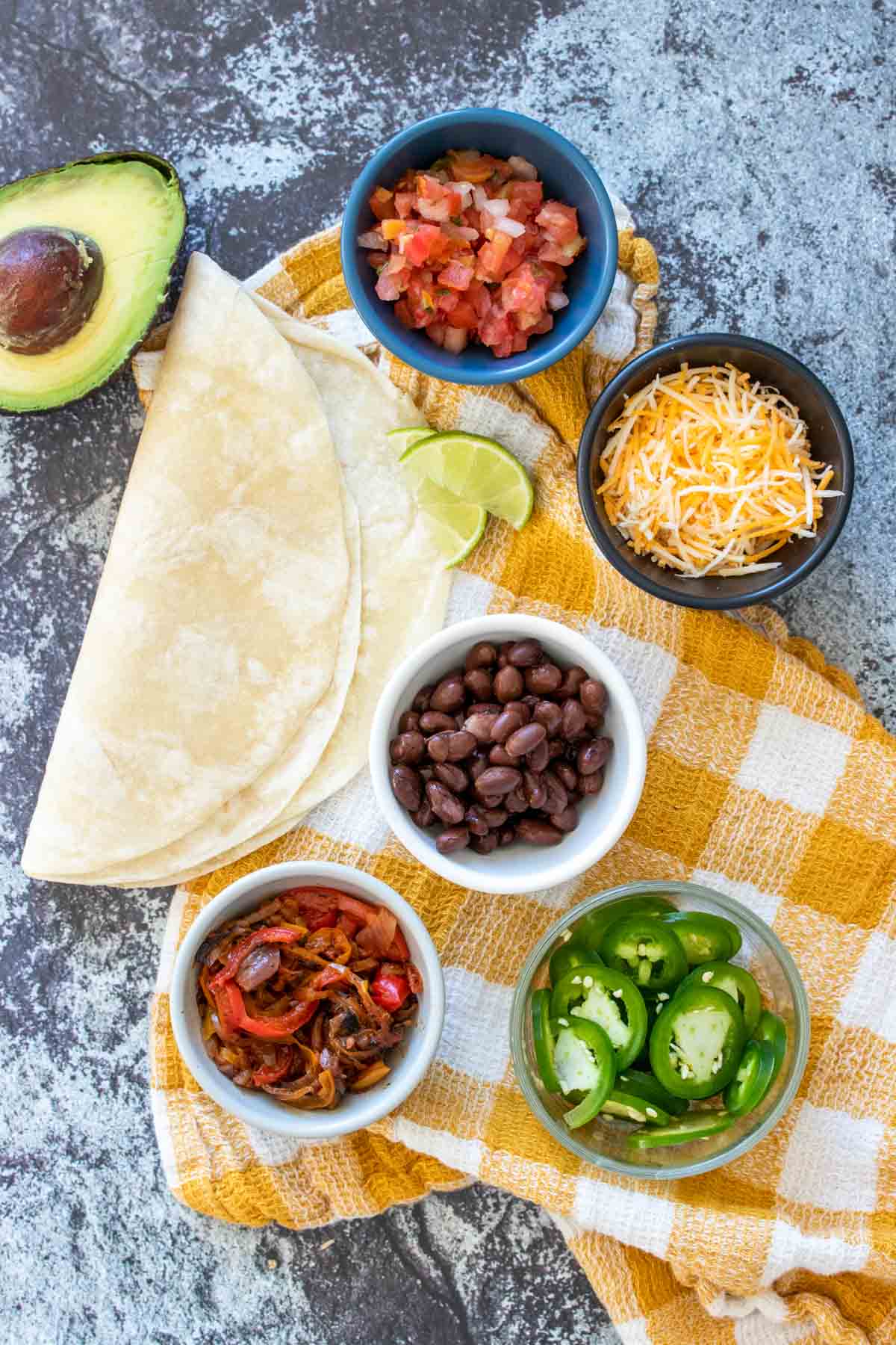 Ingredients needed to make a bean, sauteed pepper and cheese quesadilla and toppings for it sitting on a grey rock surface