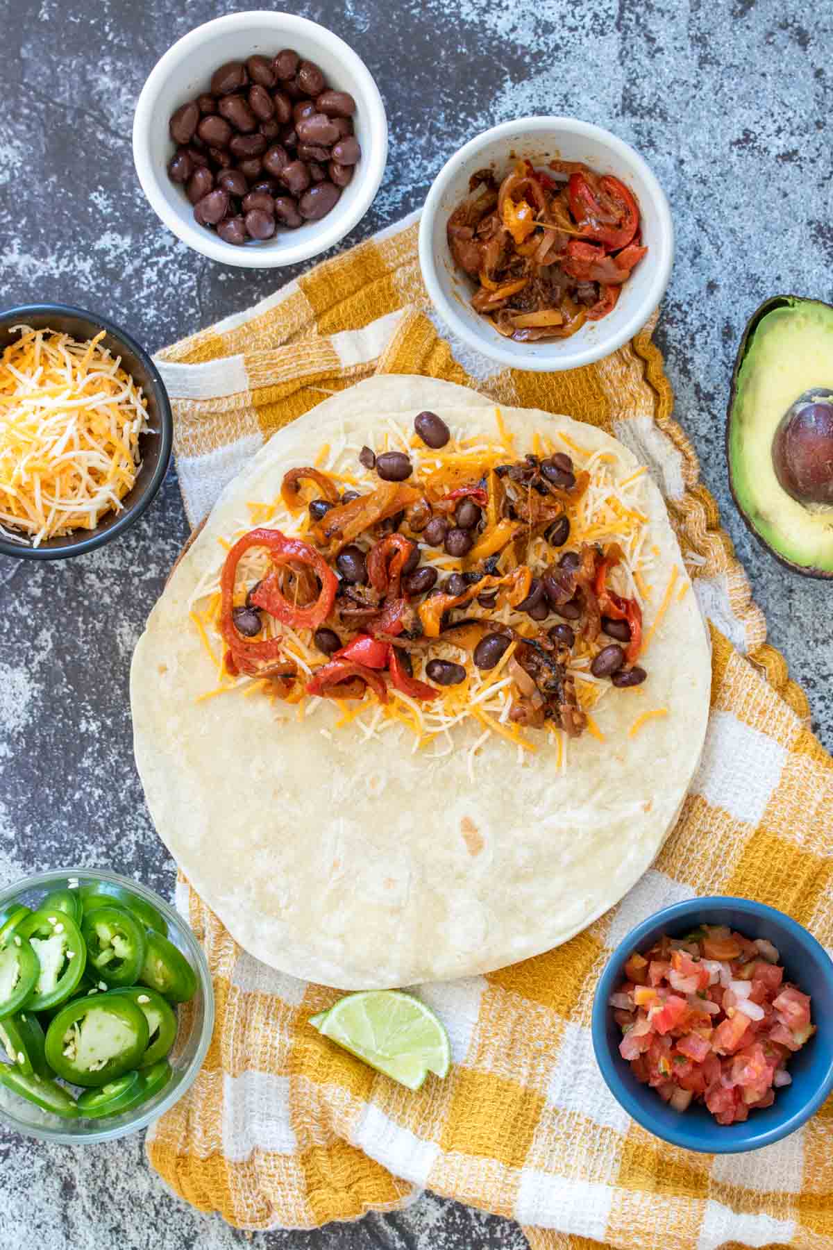 A tortilla on a yellow checkered towel with cheese, peppers and beans on half of it surrounded by toppings