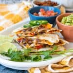 A stack of bean and pepper quesadillas on a white plate with cilantro, limes and colorful bowls.