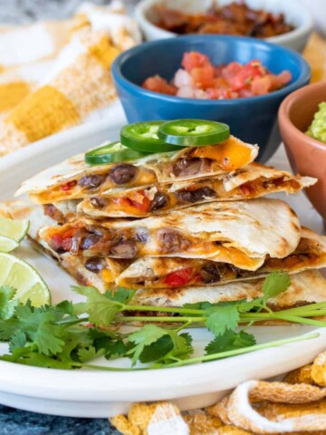 A stack of bean and pepper quesadillas on a white plate with cilantro, limes and colorful bowls.