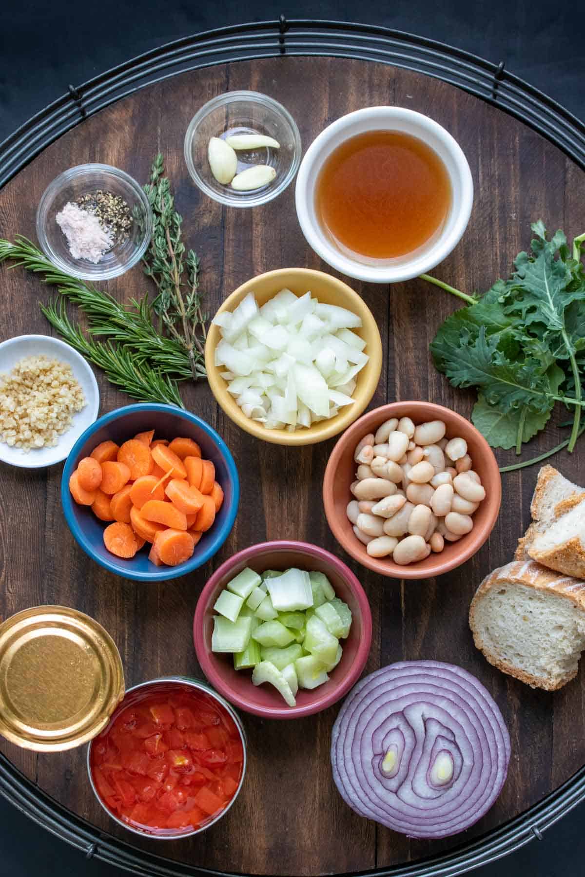 Different colored bowls on a wooden tray filled with ingredients to make a veggie ribollita soup.