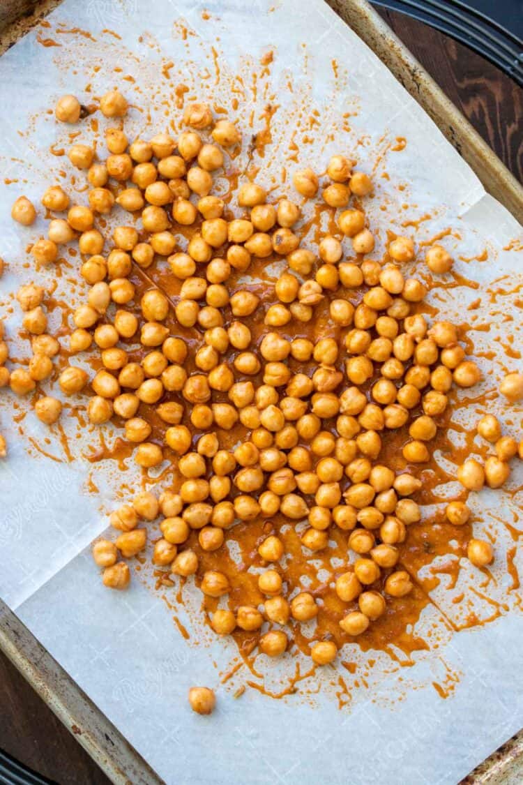 Chickpeas mixed with a dark orange colored sauce on a parchment lined baking sheet
