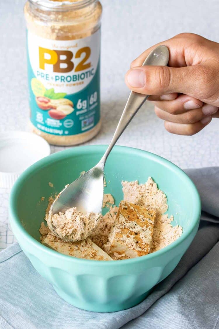 Hand using a spoon to to coat tofu pieces with a powdered mixture in a turquoise bowl