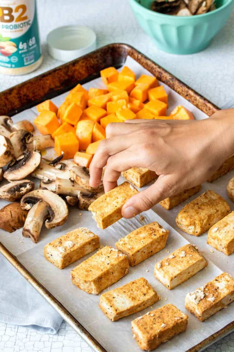 Hand putting tofu pieces on a baking sheet with veggies on it