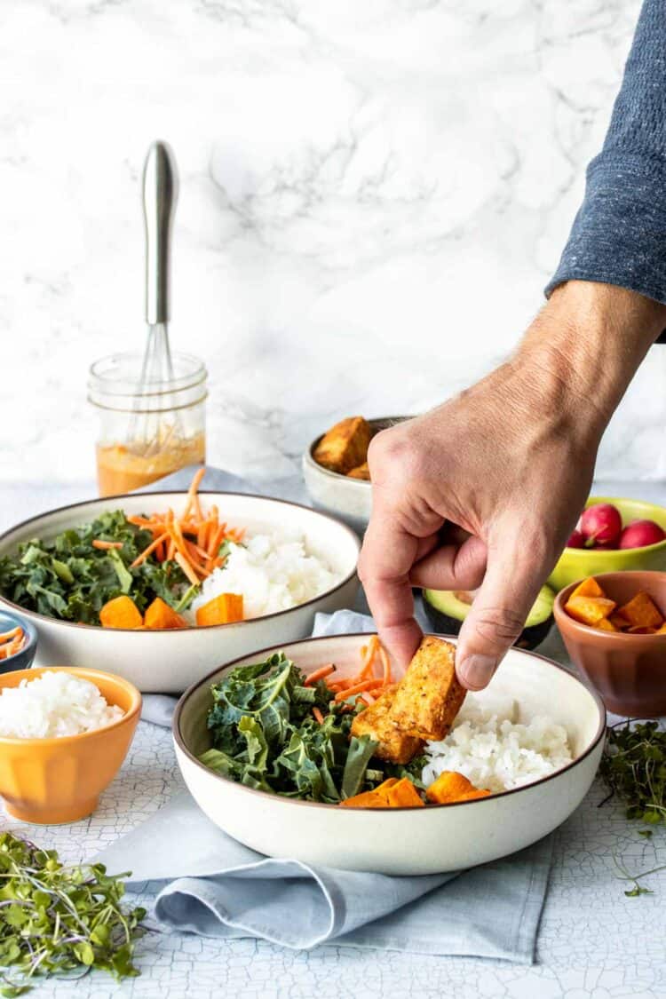 Hand putting tofu pieces in a cream colored bowl with rice, greens and potatoes