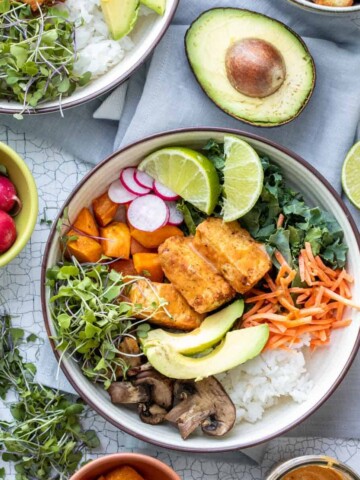 Veggies, tofu and rice in a large cream bowl surrounded by smaller bowls and toppings