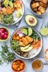 Top view of two rice bowls topped with greens, veggies, tofu and surrounded by different toppings in bowls.