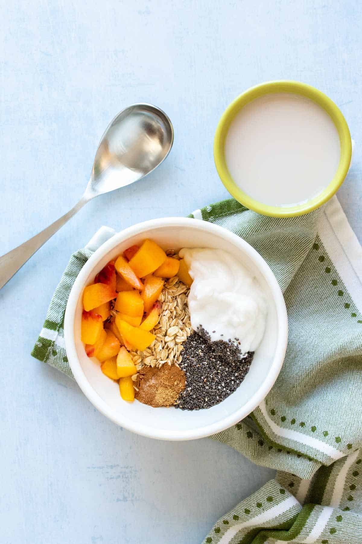 A white bowl with yogurt, peaches, chia seeds, oats and spices in it next to a spoon and milk