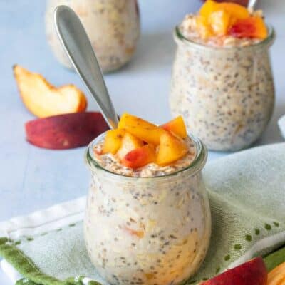 A glass jar with creamy oats and peaches on top in front of two other similar jars next to sliced peaches