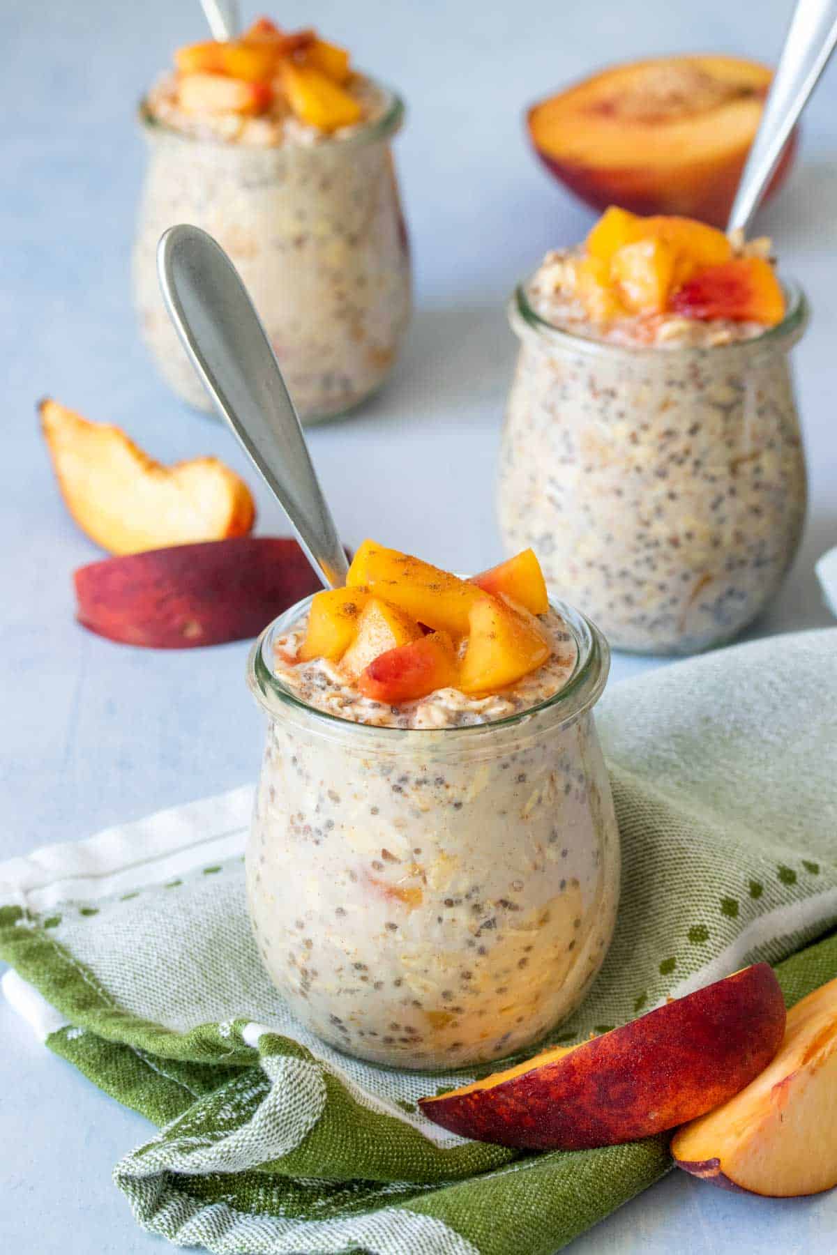 A glass jar with creamy oats and peaches on top in front of two other similar jars next to sliced peaches