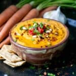 Brown bowl on a dark surface with an orange colored hummus dip topped with spices and chopped parsley.