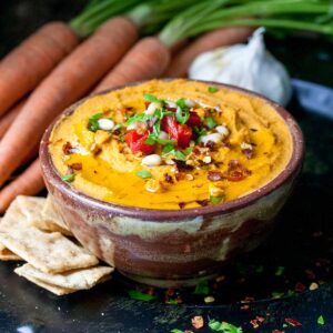 Brown bowl on a dark surface with an orange colored hummus dip topped with spices and chopped parsley.