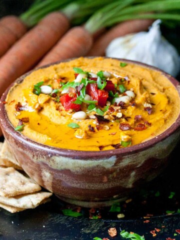 Brown bowl on a dark surface with an orange colored hummus dip topped with spices and chopped parsley.