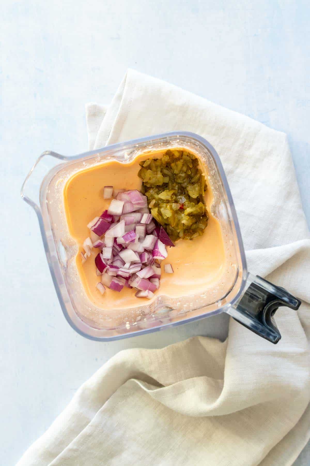 Top view of a blender with an orange creamy sauce topped with relish and chopped red onion