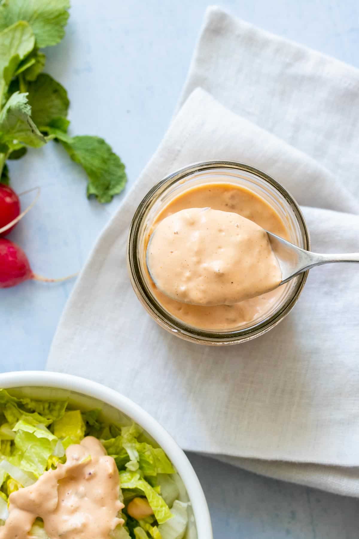 A spoon coming out of a jar with thousand island dressing on it next to a bowl of salad with dressing