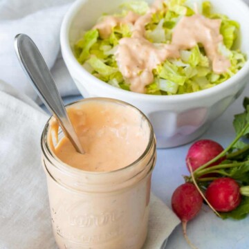 A glass jar with thousand island dressing sitting on a light colored napkin in front of a bowl of salad
