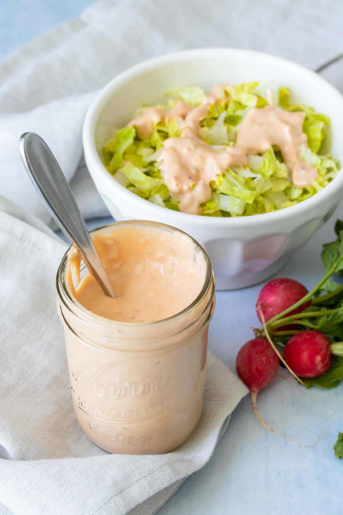 A glass jar with thousand island dressing sitting on a light colored napkin in front of a bowl of salad