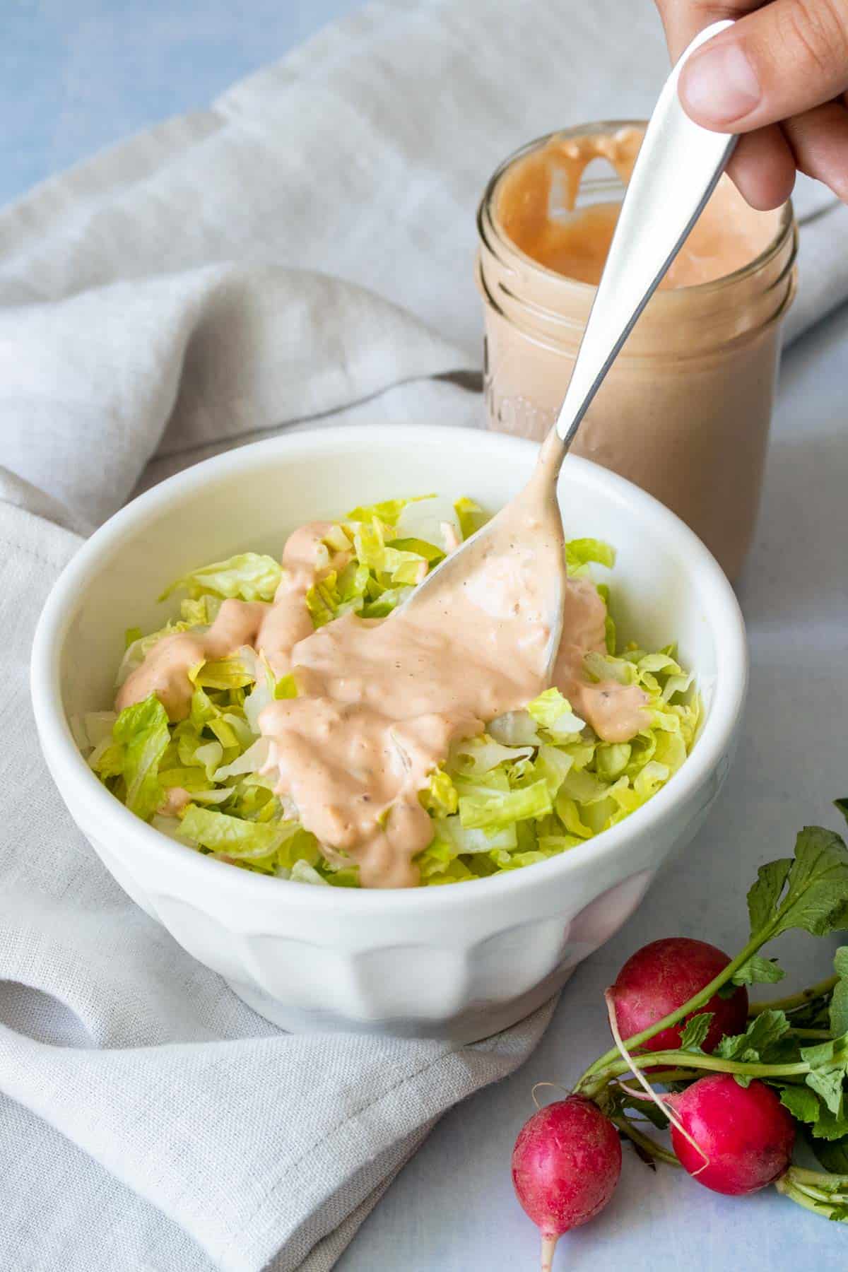 A spoon putting thousand island dressing on chopped lettuce in a white colored bowl