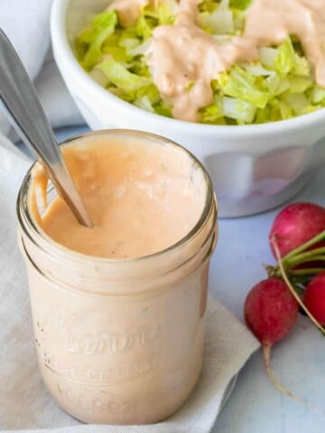 A glass jar with a creamy orange sauce and a spoon in it next to radishes and salad