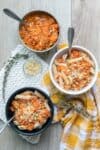 Grey bowl with red sauce, and white and black bowls with pasta and red pasta sauce on a wooden surface with towels