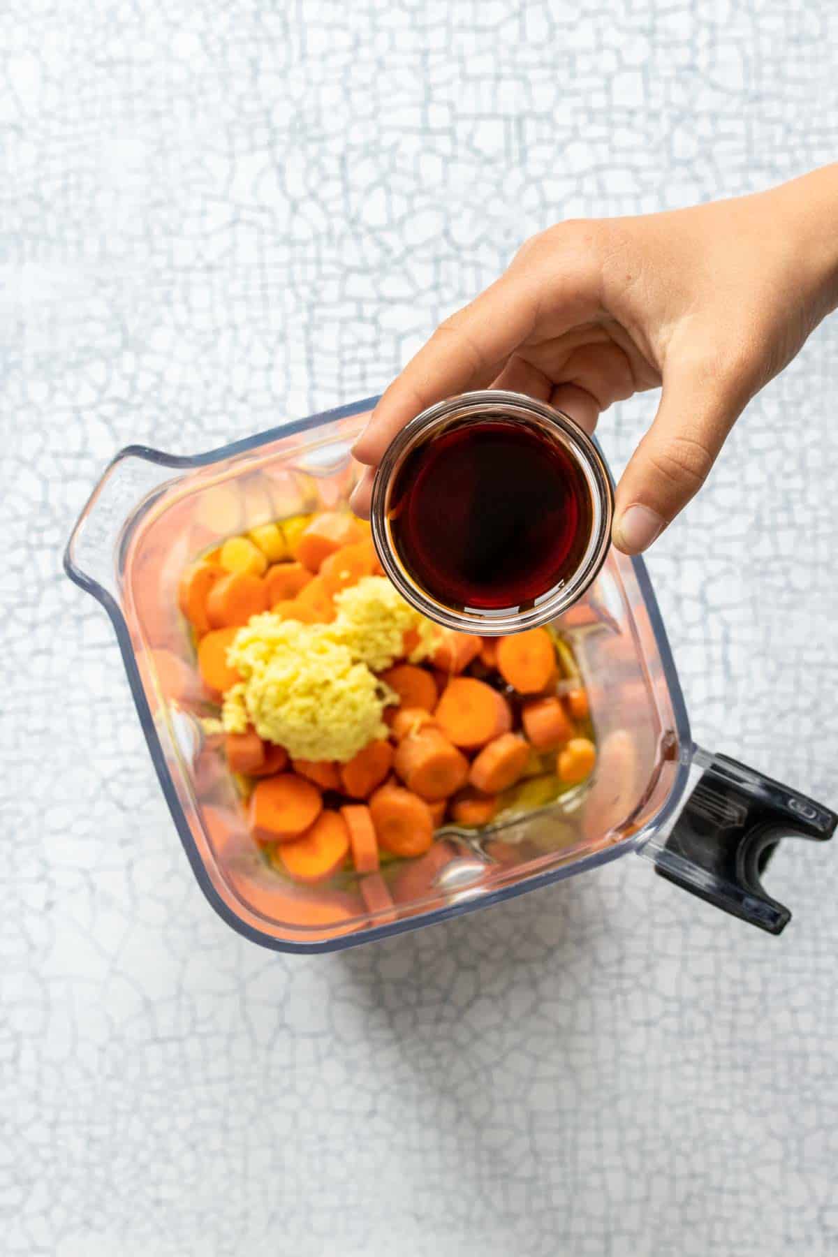 Hand holding a small glass bowl of soy sauce over a blender with carrots, onion and ginger