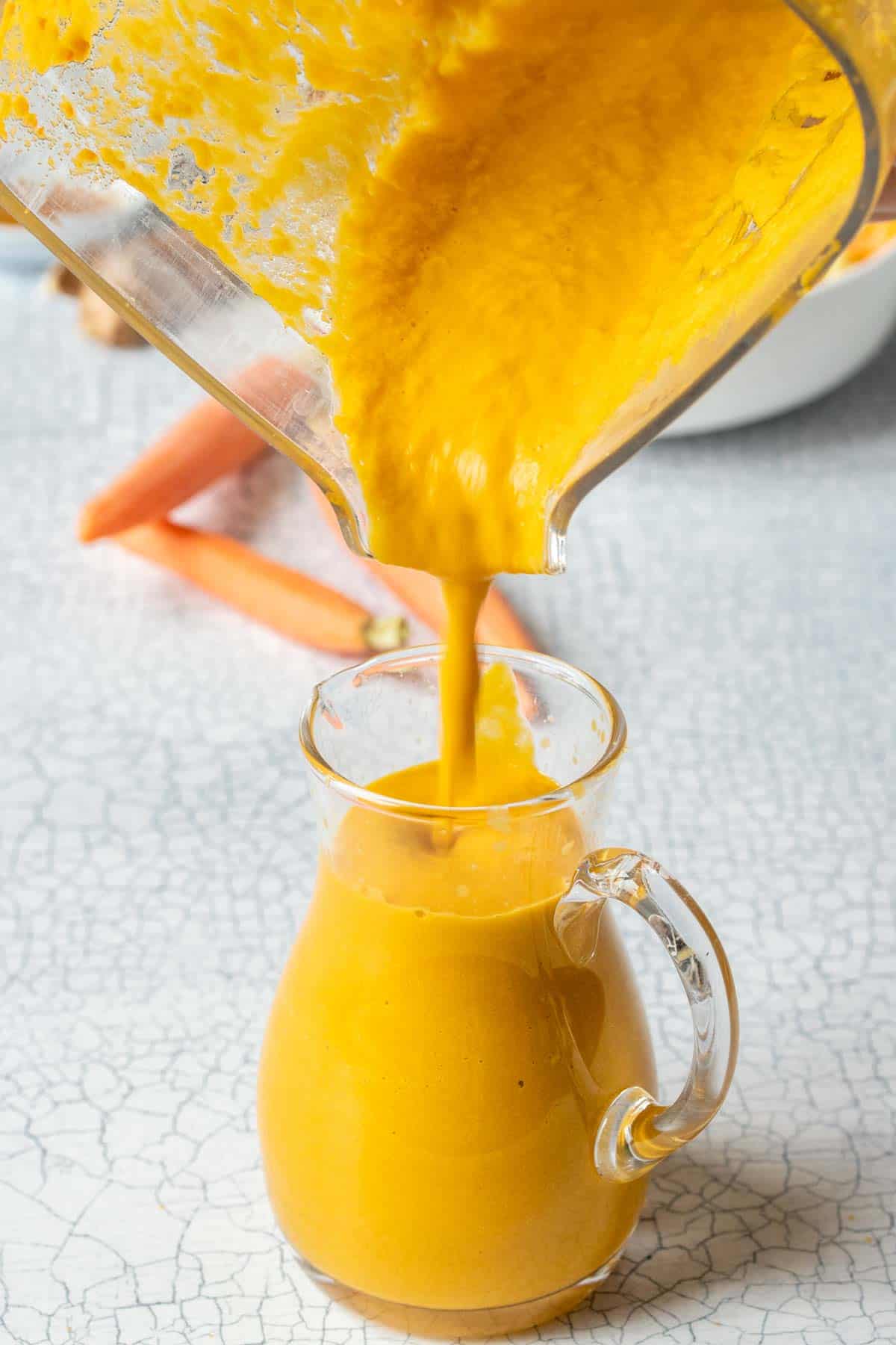 A blender pouring a vibrant orange dressing into a small glass pitcher
