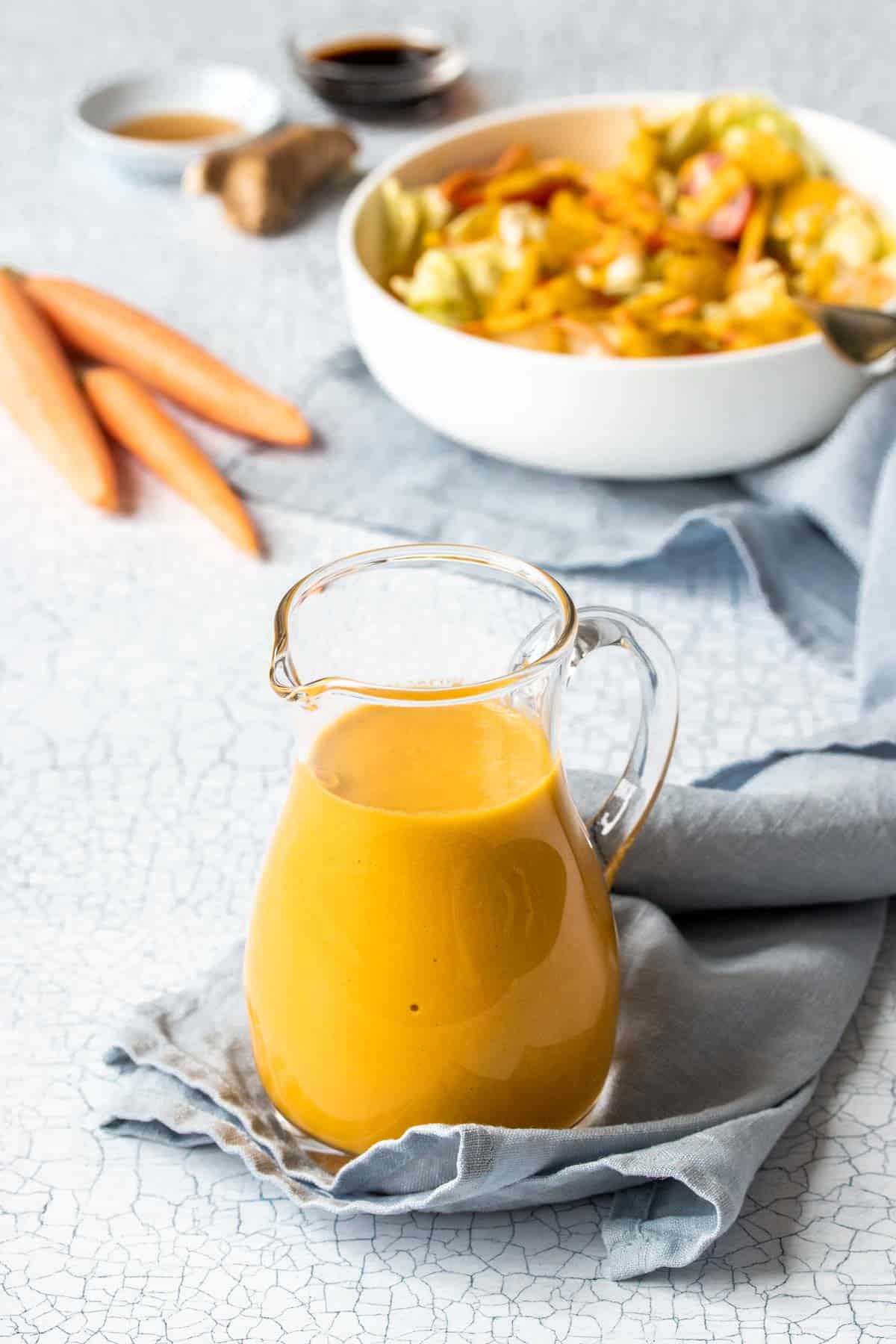 A small glass pitcher with a bright orange dressing in it in front of a white bowl of salad and carrots