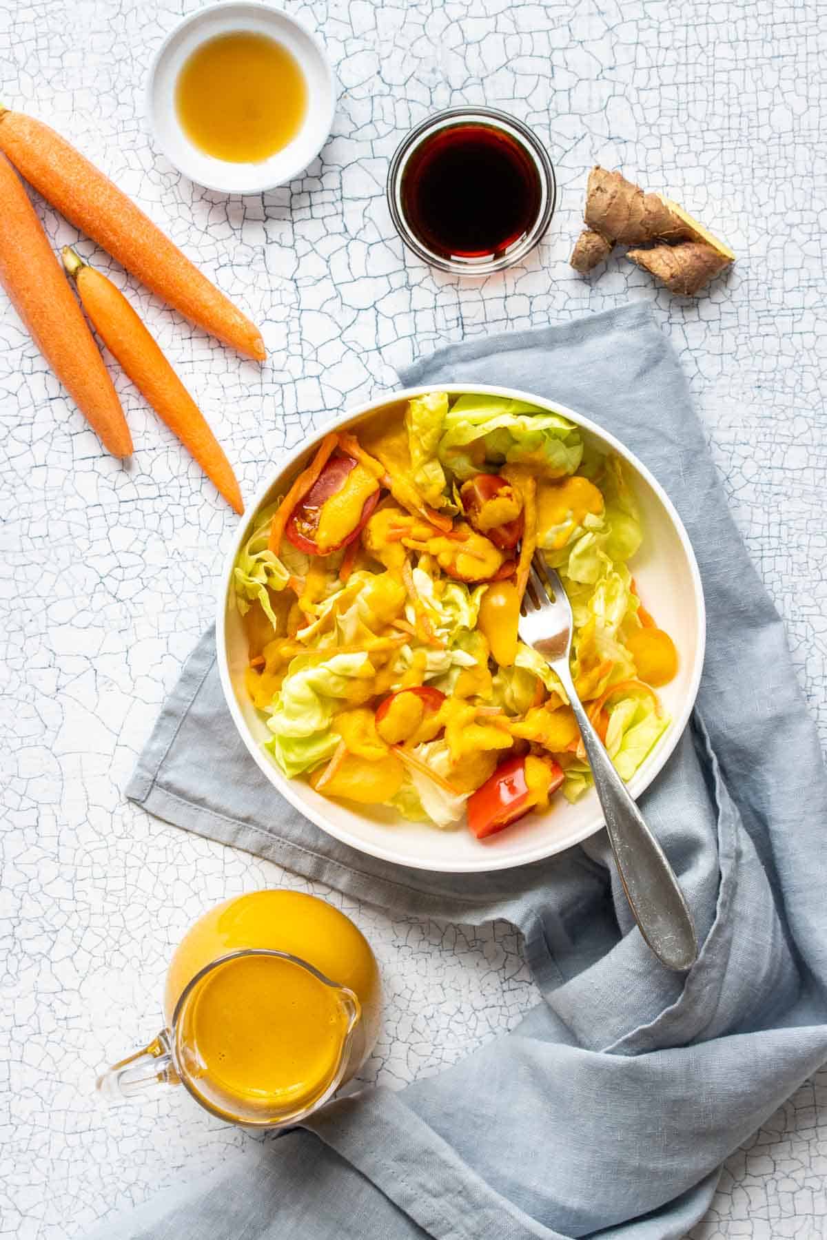 Top view of a white bowl with salad in it topped with an orange dressing and tomatoes next to a pitcher of dressing and other condiments