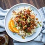 A grey plate on a grey napkin topped with rice covered with crispy orange cauliflower sprinkled with green onions.