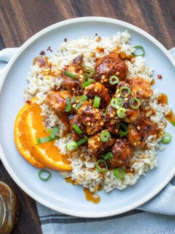 A grey plate on a grey napkin topped with rice covered with crispy orange cauliflower sprinkled with green onions.