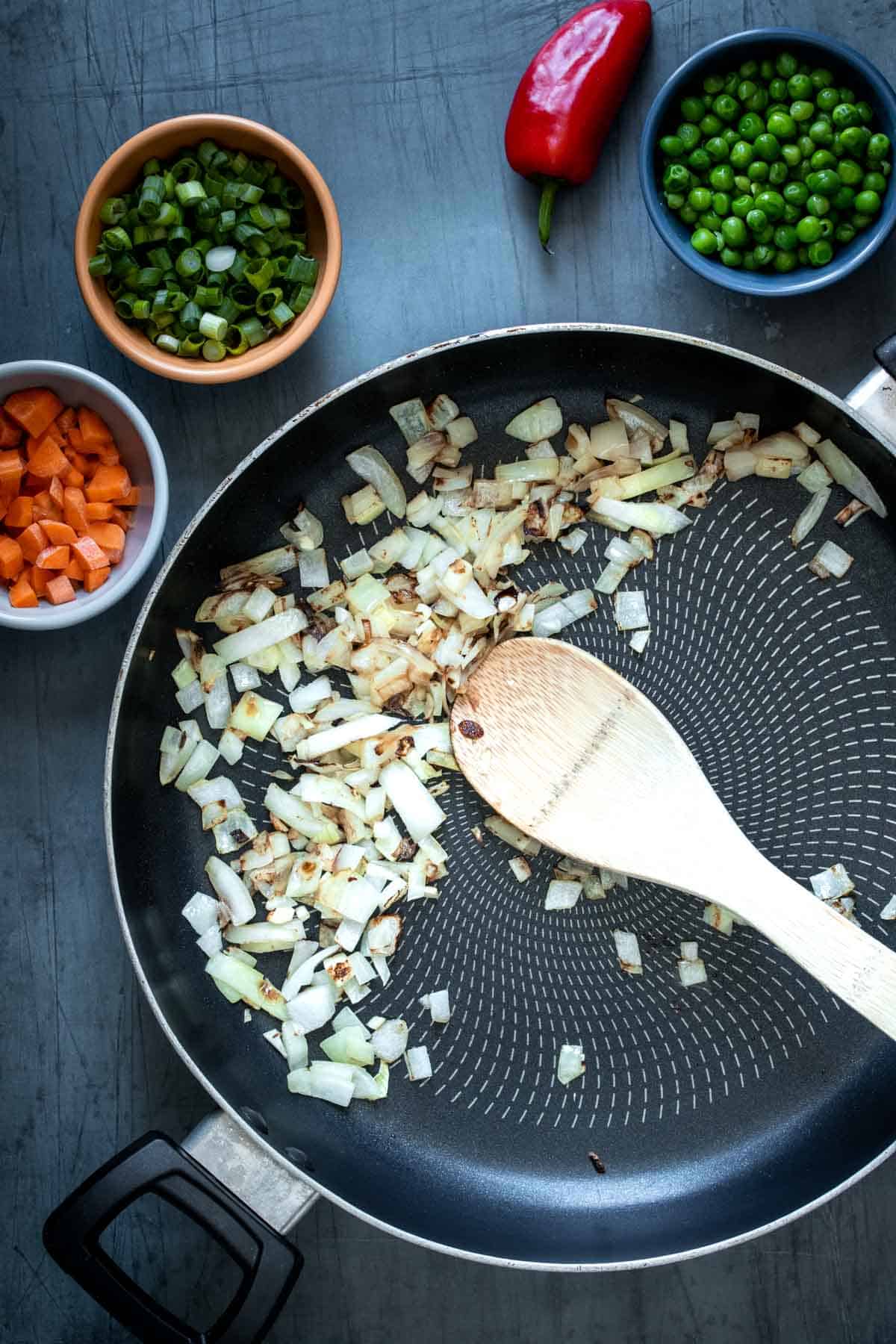 A wooden spoon mixing white onion in a pan and colorful bowls with ingredients around it