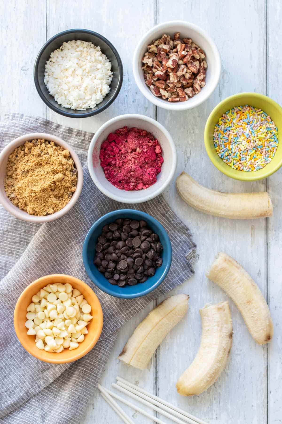 Bananas with chocolate chips and different toppings for making chocolate covered bananas in bowls on a white wood surface