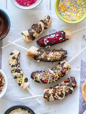 Six chocolate covered bananas covered with toppings on a piece of parchment paper