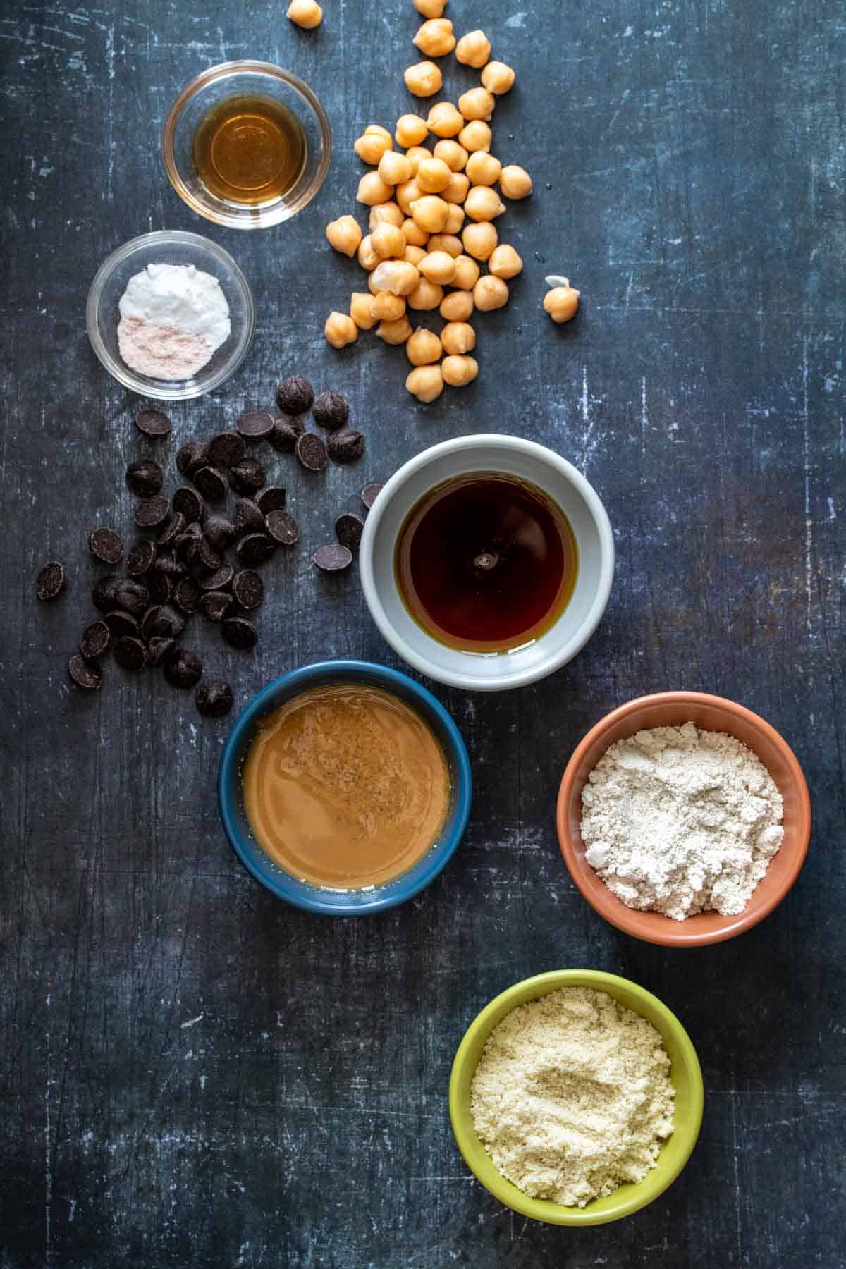 Top view of ingredients needed to make chickpea blondies vegan in bowls on a dark surface.