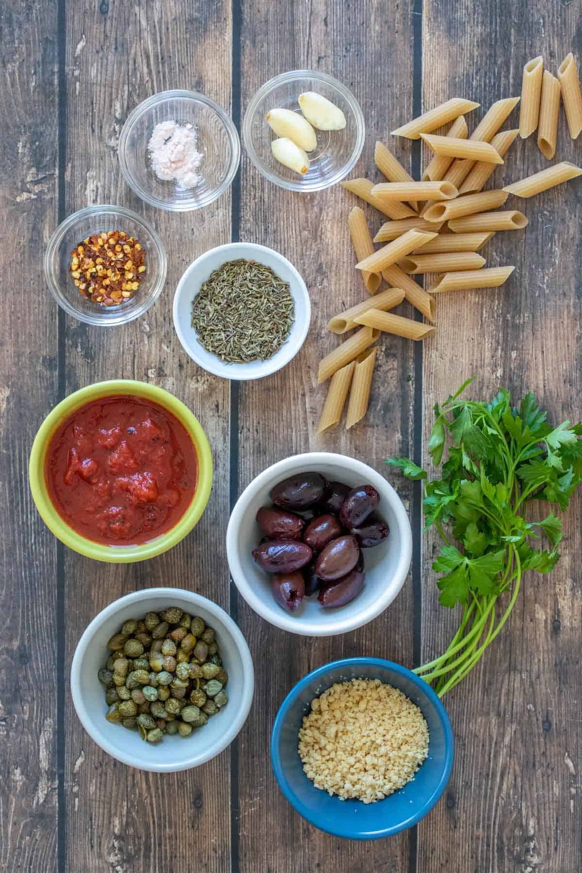 Penne pasta and parsley next to bowls of ingredients needed to make pasta puttanesca on a wooden surface.