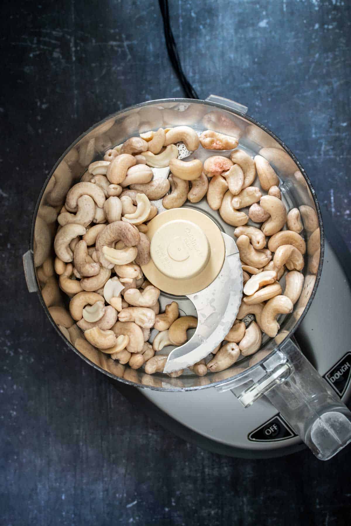 Top view of a food processor with cashews inside of it on a dark surface