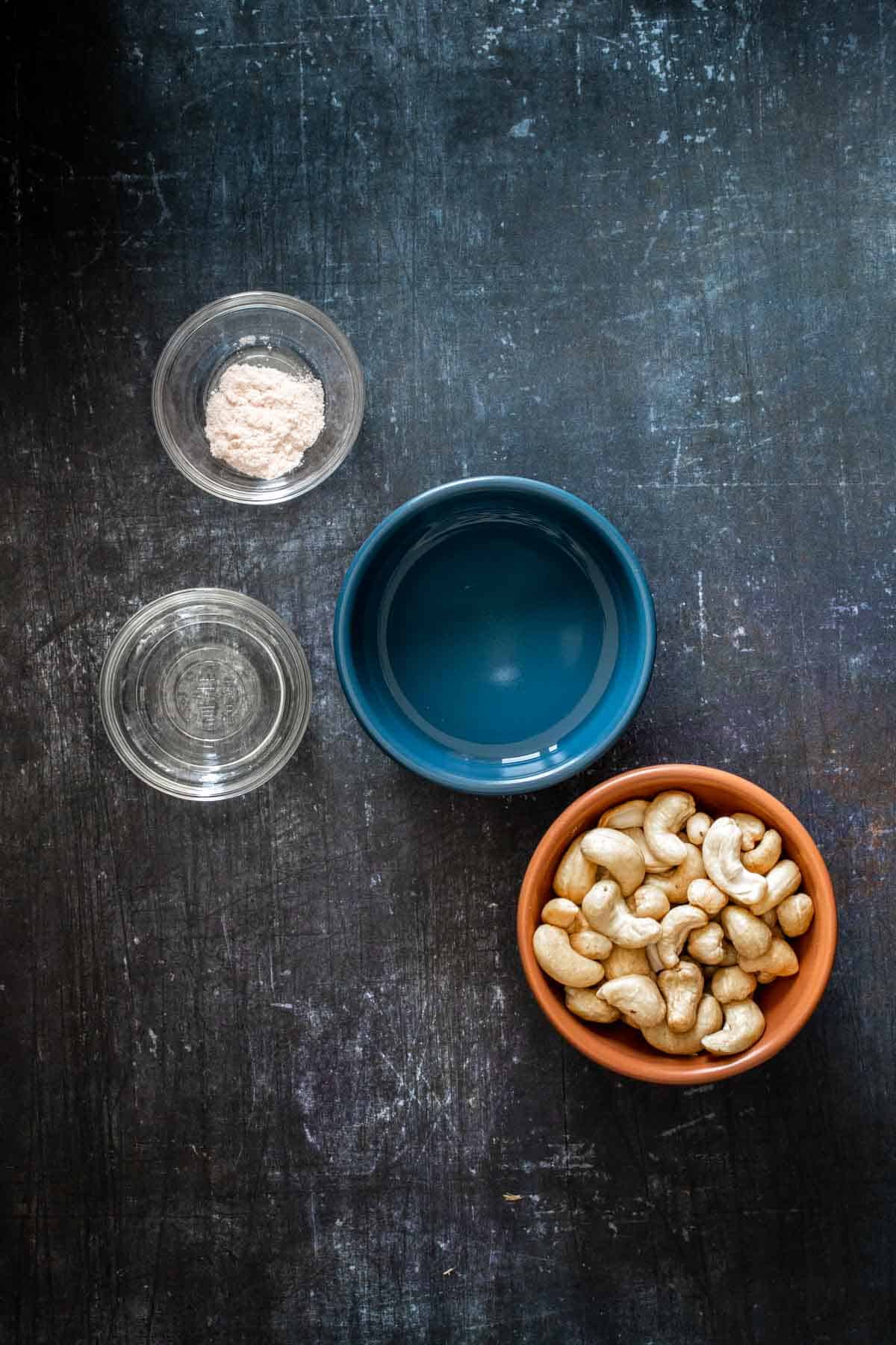 Top view of bowls with cashews, water, vinegar and salt on a dark surface