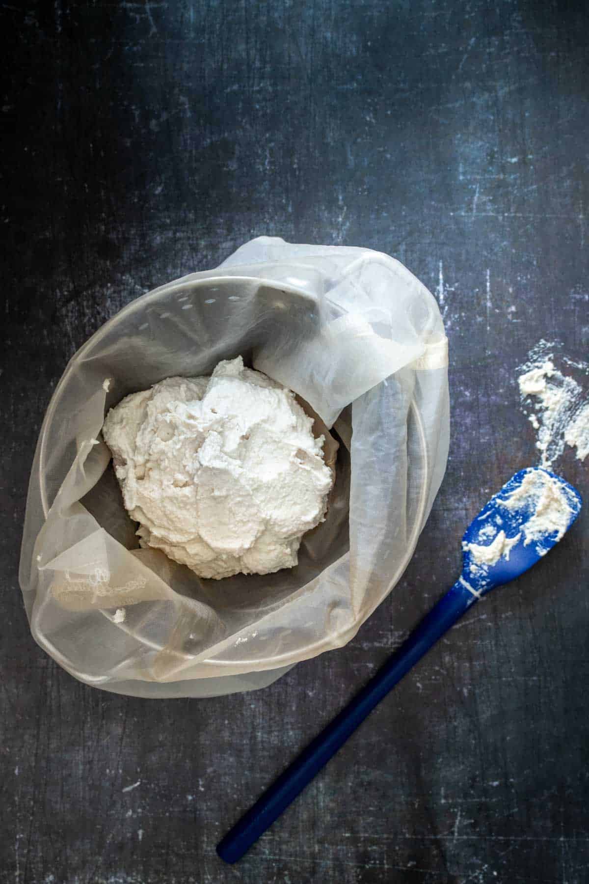 Top view of a thick creamy mixture in a cheese cloth over a strainer with a blue spatula next to it