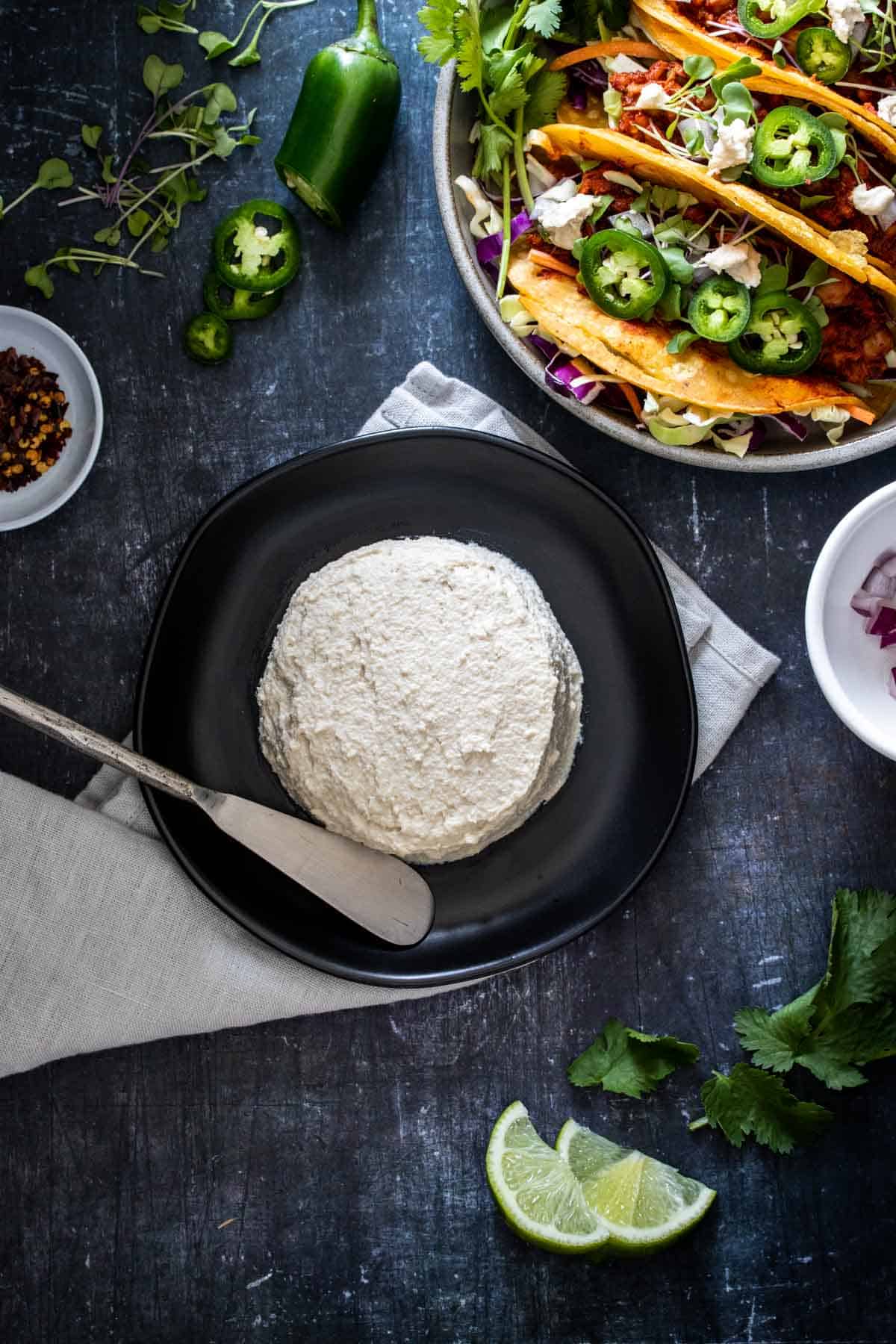 Top view of a wheel of creamy cheese on a black plate next to a plate of tacos with the cheese on them