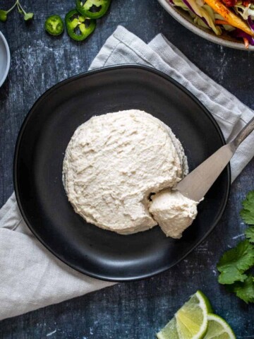 A round shape of thick creamy cheese with a knife taking some from it on a black plate.