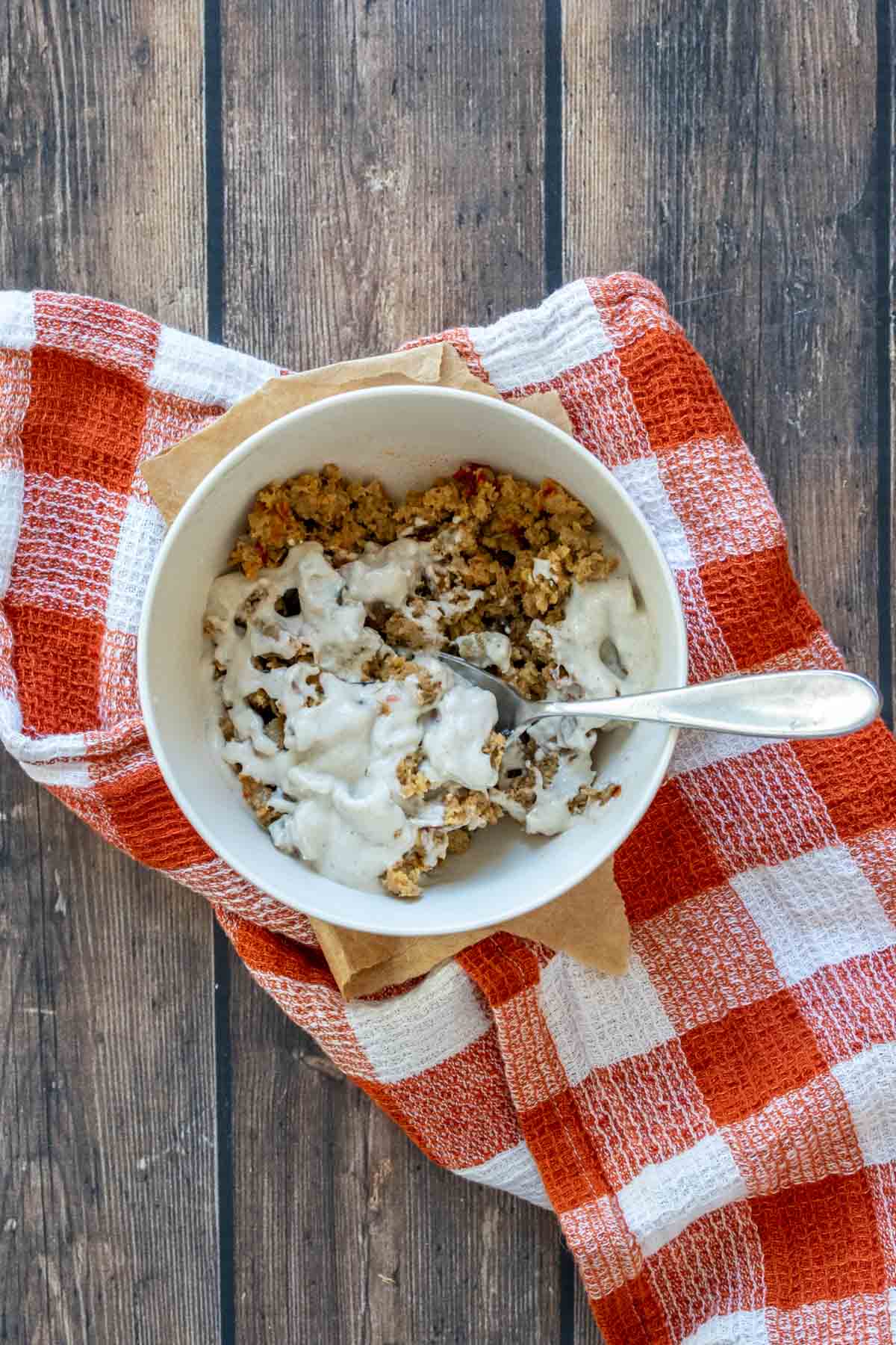 A spoon mixing crumbled sausage and a creamy sauce in a cream colored bowl on a dark orange checkered towel