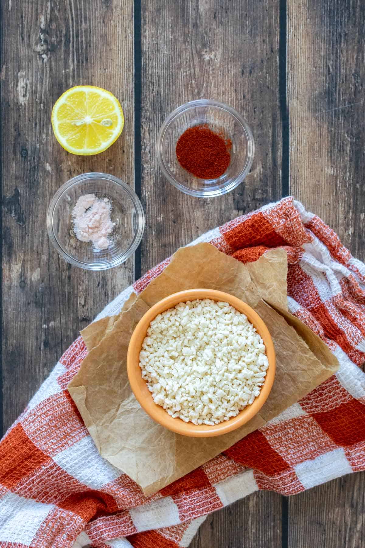 A bowl of breadcrumbs, salt, paprika and a lemon on a wooden surface with a checkered orange towel