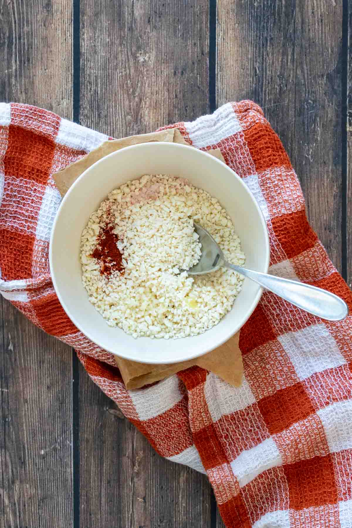 A cream bowl with a spoon in it and breadcrumbs and spices on a dark orange checkered towel