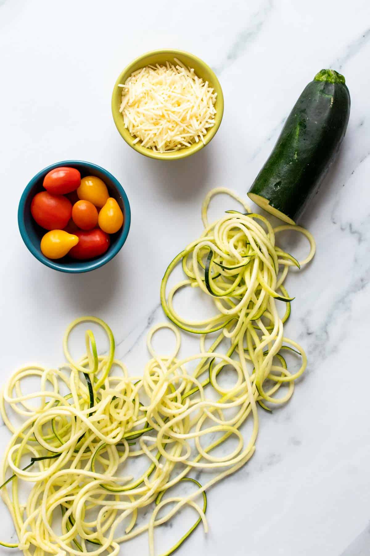 Top view of a zucchini with spiral zucchini noodles coming from it next to parmesan and tomatoes