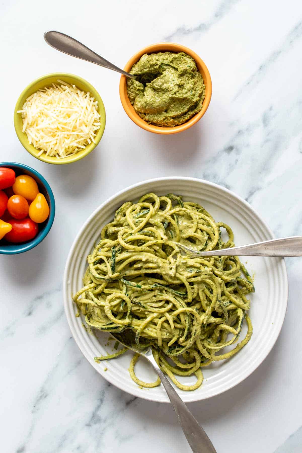 A white plate with zucchini noodles mixed with pesto and bowls of pesto, tomatoes and parmesan next to it